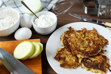 zucchini fritters on a white plate, sour cream on a brown wooden background, a knife, eggs, pieces...