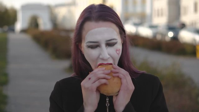 Mime girl in a black coat eats burger on the street.