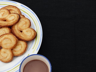 Heart-shaped puff pastry (palmeritas) with a blue cup of chocolate with a black background.(negative space)