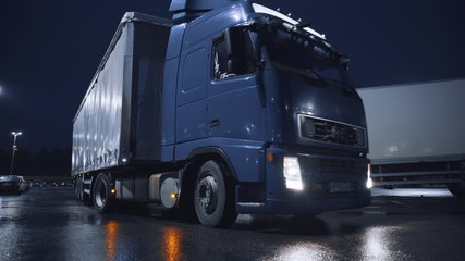 Blue Semi-Truck with Cargo Trailer Drives Off From Overnight Parking Space where Other Trucks are Standing. Long Haul Truck Leaves Parking Lot, Transporting Cargo / Goods Across Continent. Rainy Night