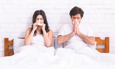 Young married couple sneezing into paper napkins in bed