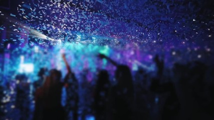 people jump with raised hands under white confetti fall against blurry illuminated stage in night club slow motion