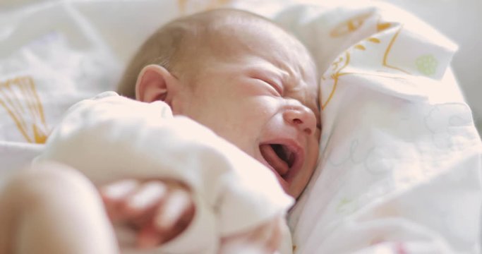 Portrait of sleepy sad newborn baby is crying in bed with closed eyes. Angry upset baby shouting and grimacing face needing in parent's attention and care. Concept of babyhood and newborn care.