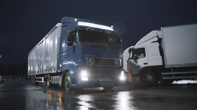 Blue Semi-Truck with Cargo Trailer Drives Off From Overnight Parking Space where Other Trucks are Standing. Long Haul Truck Leaves Parking Lot, Transporting Cargo / Goods Across Continent. Rainy Night
