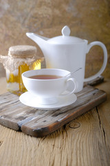 A white cup of hot tea and a white kettle of honey on a wooden table.