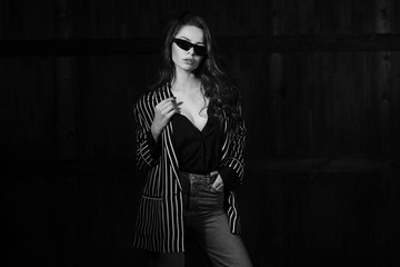 Studio portrait of fashionable young woman in male style clothes posing against dark background. Straight hair, sunglasses, grey jeans, black blouse and striped jacket. Vogue style.