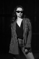 Naklejka premium Body length interior portrait of stylish young female modelwearing grey jeans and striped blazer standing and posing in dark room. Vogue style. Studio shot