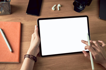 hands using tablet with blank screen and holding digital pen, smartphone with camera and accessories on wood table.