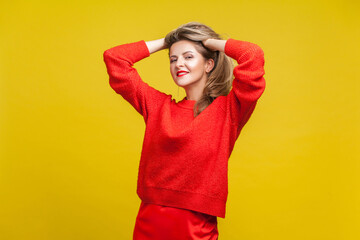 Portrait of impressive happy blonde woman with red lipstick in bright sweater standing with hands in hair, looking at camera with seductive smile. indoor studio shot isolated on yellow background