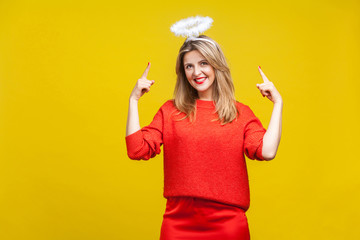 This is me angel! Portrait of proud adorable young woman with bright makeup in red casual clothes standing, pointing at hola on her head and smiling. indoor studio shot isolated on yellow background © khosrork