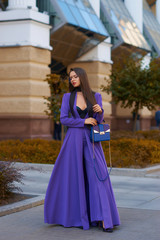 Fototapeta premium Young beautiful caucasian woman with long straight hair and makeup in purple trousers and long coat cape standing and posing at city street in autumn