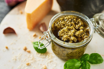 Pesto sauce or pesto genovese in a glass jar with pine nuts, parmesan, basil, oil and garlic on white marble cutting board. Copy space.