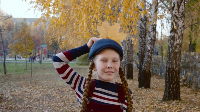 Girl teenager in striped clothes and blue cap with yellow maple leaf near head like crow. Portrait happy girl with autumn foliage in autumn park. Beautiful model posing with autumn leaf in park