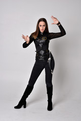full length portrait of a pretty brunette woman wearing black leather fantasy costume with a gun. standing pose on a studio background.