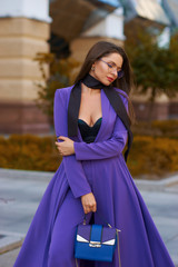 Young beautiful caucasian woman with long straight hair and makeup in purple trousers and long coat cape standing and posing at city street in autumn