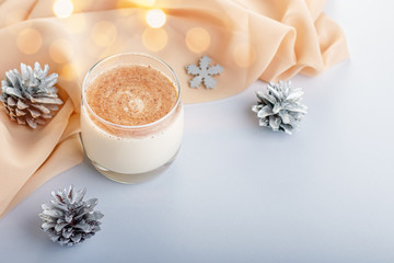 Traditional Christmas cocktail Eggnog with eggs, alcohol, grated nutmeg and cinnamon closeup, copy space. Sweet traditional drink on grey table with beige decorations, burning lanterns and pine cones