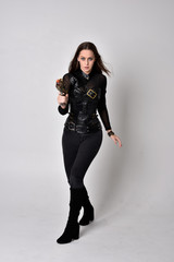 full length portrait of a pretty brunette woman wearing black leather fantasy costume with a gun. standing pose on a studio background.