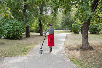 A young woman with purple hair rides an electric scooter in a park. A stylish girl with a shaved...