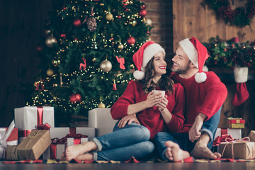 Photo of dreamy couple newyear night in decorated garland lights room sitting cozy on floor near x-mas tree drinking hot beverage indoors wearing red sweaters and santa hats