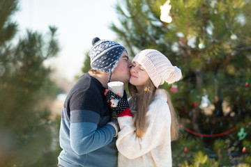 The guy with the girl walk and kiss in the winter forest with a mug of hot drink. A cozy winter walk through the woods with a hot drink. Loving couple, winter holidays