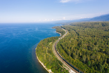 Aerial view of a freight train on the railroad of Trans-Siberian Railway on the shore of Baikal Lake with green forest trees in a sunny summer day. East Siberian Railway in Buryatia, Siberia, Russia