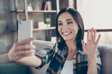 Photo of charming lady holding telephone making selfies speaking skype waving palm saying hi sitting comfort sofa wear casual checkered shirt apartment indoors © deagreez