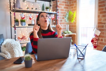 Beautiful woman sitting at the table working with laptop at home around christmas lights amazed and surprised looking up and pointing with fingers and raised arms.