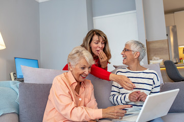 Portrait of 3 senior female friends. Group of senior female friends sitting together in living room and watching movie on on laptop. Home party. Modern happy grandmothers