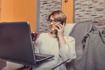 Middle aged woman talking on phone and working on laptop at home