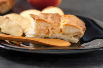 Slice Charlotte apple pie on a black plate with a wooden spoon on a black wooden table.Close up.