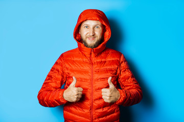 Man in a red jacket with a hood makes a gesture with his hands on a blue background. Concept of winter, autumn, cold