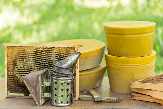 Large round pieces of natural beeswax on the background of beehives in the garden. Large round pieces of beeswax, bee smoker and beekeeper's tool on the background of beehives in the garden. - Powered by Adobe