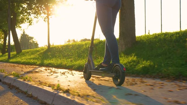 SLOW MOTION, LENS FLARE, CLOSE UP, LOW ANGLE, COPY SPACE: Cinematic shot of an unrecognizable modern girl wearing jeans riding an electric scooter down the scenic fall colored avenue at golden sunset.