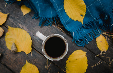 On a old wooden table in the autumn park is a cup of tea-coffee. A blue warm plaid scarf is...