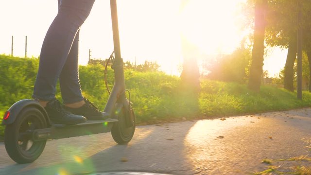 SLOW MOTION, SUN FLARE, COPY SPACE, LOW ANGLE Cinematic shot of a modern woman in jeans riding an e-scooter down the empty pavement at sunset. Unrecognizable girl cruises on e-scooter on sunny evening