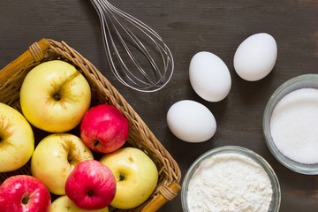 Top view of apples, eggs, sugar, flour and whisk on wooden background