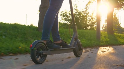 COPY SPACE, CLOSE UP, LOW ANGLE: Young woman in jeans rides an electric scooter along sidewalk at beautiful sunrise. Unrecognizable girl enjoys a scenic e-scooter ride along an autumn colored avenue. - Powered by Adobe