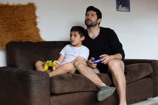 Padre e hijo latinoamericanos juegan video juegos sentados en el sof&aacute; en &eacute;poca de cuarentena