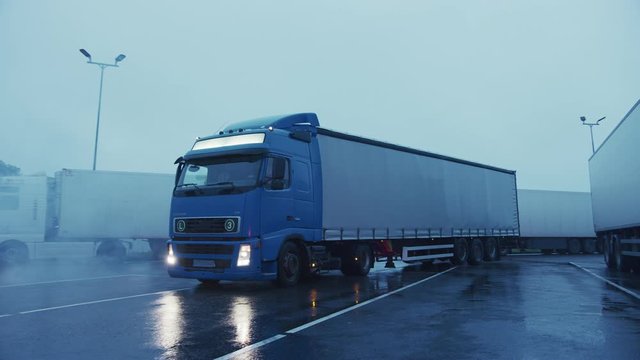 Blue Semi-Truck with Cargo Trailer Drives Into Parking Lot where Other Trucks are Standing. Industrial Logistics Area. Loading or Unloading Space for Long Haul Trucks. Side View Arc Shot
