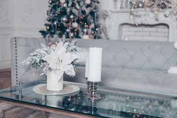 Classic white Christmas interior with modern elements and New Year tree decorated. Fireplace with grey sofa and glass table