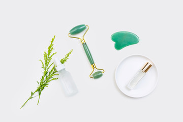 Gua sha stone, jade roller, clear perfume, white glass eye dropper serum bottle / still life arranged with green wildflowers and isolated on white surface / natural clean beauty concept