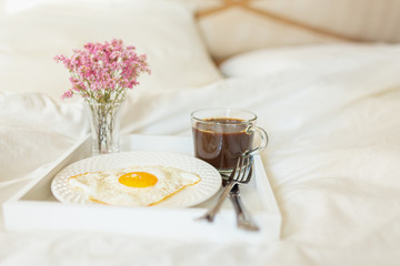 White tray with breakfast on a bed in a hotel room. Fried egg, cup of coffee and flowers in white sheets in light bedroom.