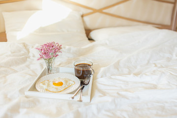 White tray with breakfast on a bed in a hotel room. Fried egg, cup of coffee and flowers in white...