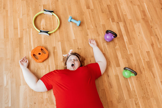 Tired fat girl dressed in the sportswear and with a bandage on her head is laying on the floor next to the sports equipment