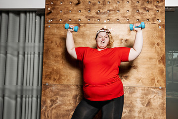 Funny fat girl dressed in the sportswear and with a bandage on her head works out with dumbbells on...