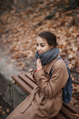 Autumn outdoor portrait of beautiful young woman in autumn park, sitting on a bench and wraps in warm knitted scarf..