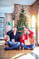 Obraz premium Young couple wearing santa claus hat sitting on the floor around christmas tree at home doing ok gesture with hand smiling, eye looking through fingers with happy face.