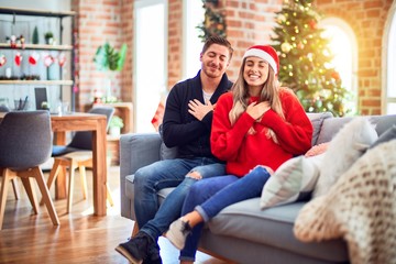 Young couple wearing santa claus hat sitting on the sofa around christmas tree at home smiling with hands on chest with closed eyes and grateful gesture on face. Health concept.