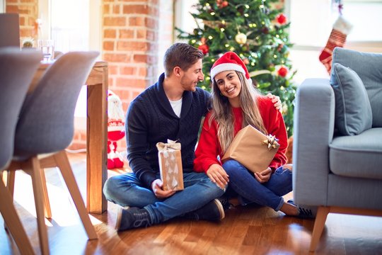 Young beautiful couple smiling happy and confident. Sitting on the floor holding gifts and hugging around christmas tree at home