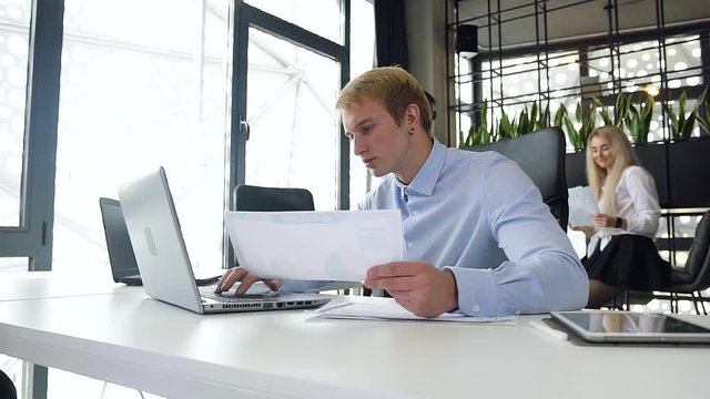 Attractive portrait of handsome young blond businessman analising report with charts with datas on laptop at his workplace in the office room